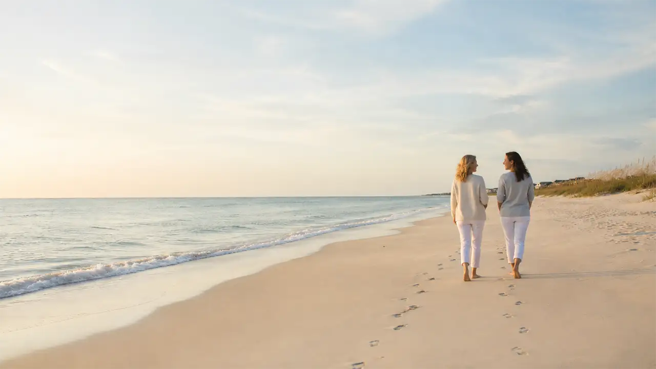 Two women walking and talking along a peaceful beach shoreline representing telehealth and walk and talk therapy at Beach Cottage Counseling.